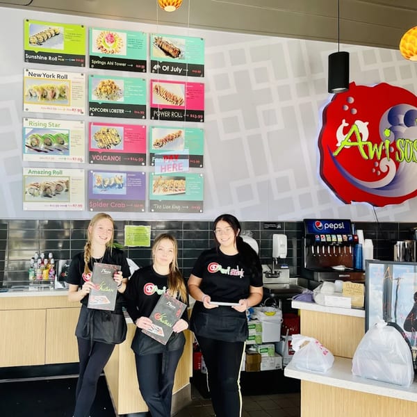 Three Awi Sushi employees stand smiling in front of a menu board and soda fountain.